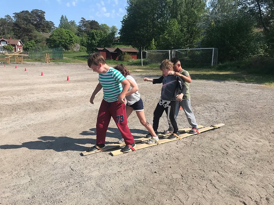 Några barn från en skolklass tävlar i en teambyggande aktivitet. Fyra barn stå på två träskidor och försöker ta sig fram mot mål.